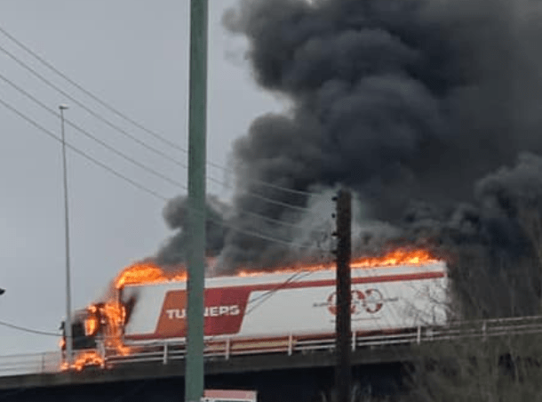 Lorry engulfed in flames on bridge, heavy smoke.